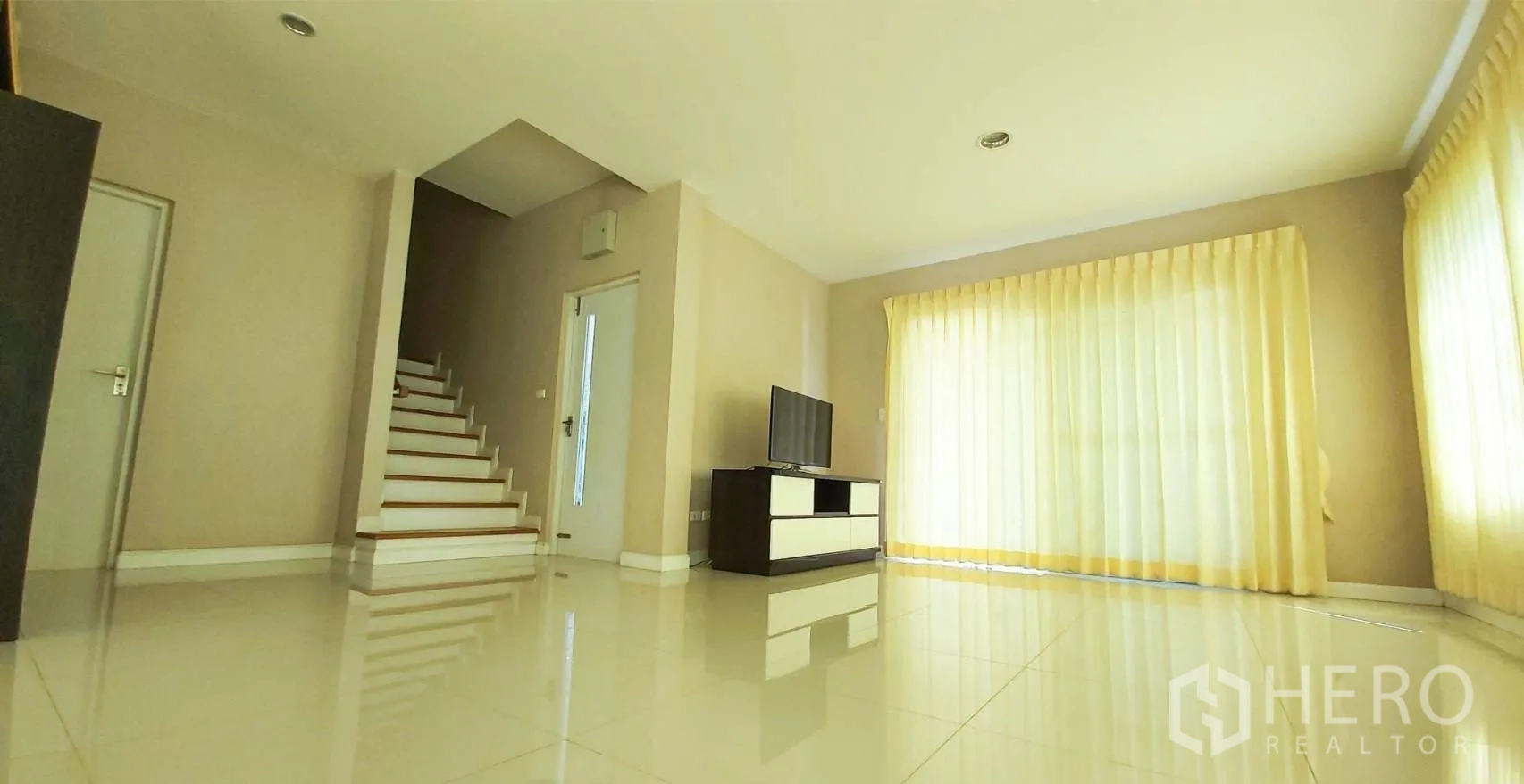 บ้าน for sale in Bang Kapi, Bangkok - มุมห้องนั่งเล่นมองไปยังบันไดและชั้นวางทีวี พร้อมม่านสีครีม