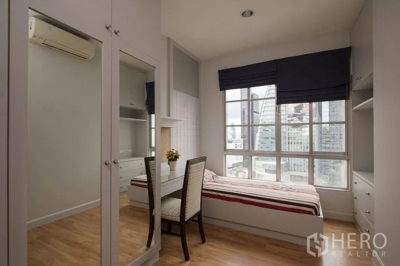 Condominium for rent in Khlong Toei, Bangkok - Corner bedroom with single bed, built-in desk and storage, and city view through large windows.