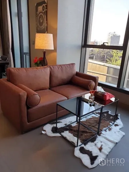 Condominium for rent in Huai Khwang, Bangkok - Living room with brown sofa, glass coffee table, floor lamp, and city view through wide windows.