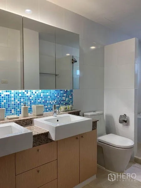 公寓 出租或出售在Phra Khanong, Bangkok - 现代卫浴，设双台盆、大镜子与坐便器，蓝色马赛克背墙。