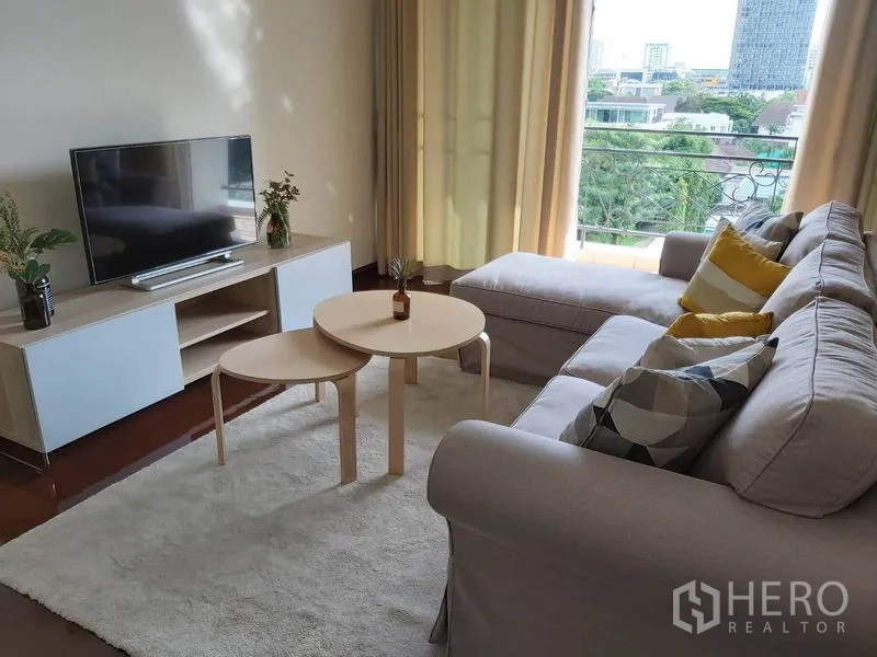 公寓 出租在Watthana, Bangkok - 明亮客厅配L形沙发、电视柜和可观绿意与天际线的阳台。