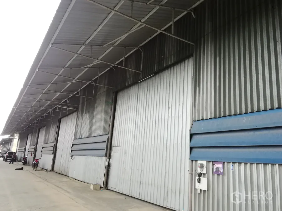 คลังสินค้า ให้เช่าในLam Luk Ka, Pathum Thani - ด้านหน้าโกดังผนังเมทัลชีท มีหลังคากันสาดยาวและประตูบานสไลด์หลายช่องในโซนอุตสาหกรรม.