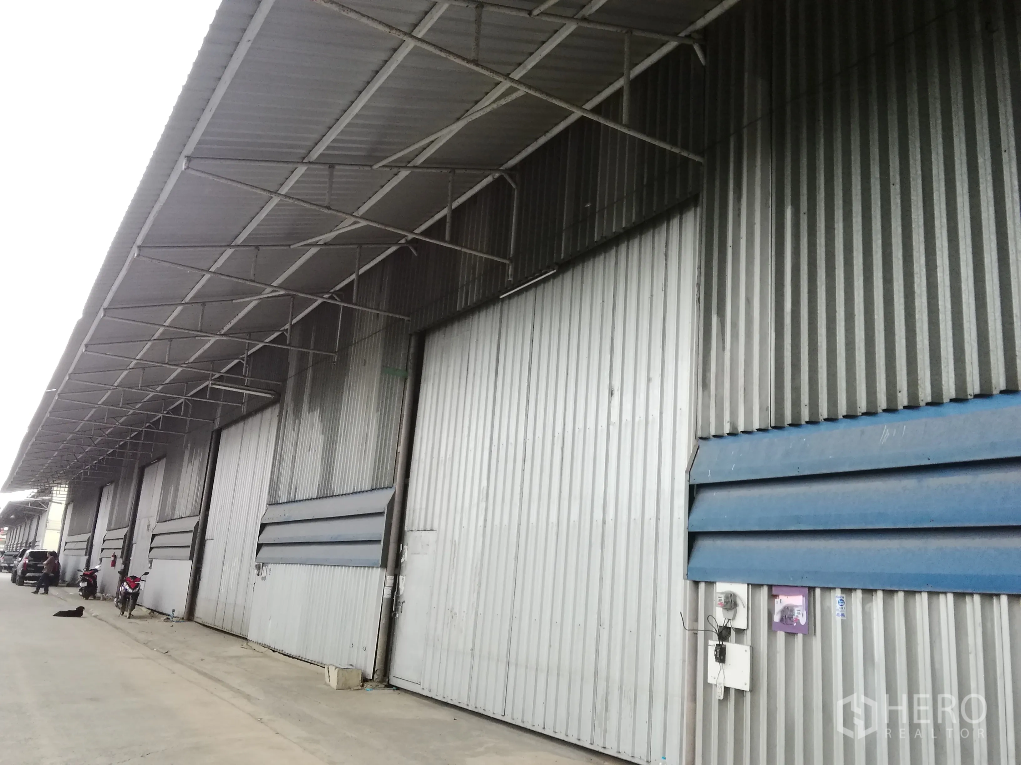 คลังสินค้า ให้เช่าในLam Luk Ka, Pathum Thani - ด้านหน้าโกดังผนังเมทัลชีท มีหลังคากันสาดยาวและประตูบานสไลด์หลายช่องในโซนอุตสาหกรรม.