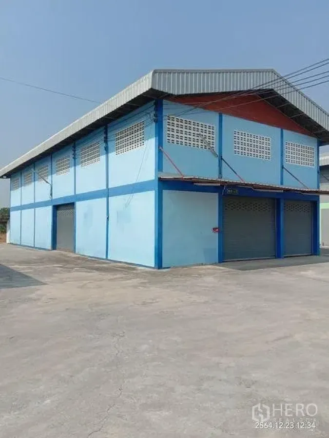Factory for rent in Lat Lum Kaeo, Pathum Thani - Corner view of a blue factory building with metal roof and multiple rolling shutter doors on a paved yard.