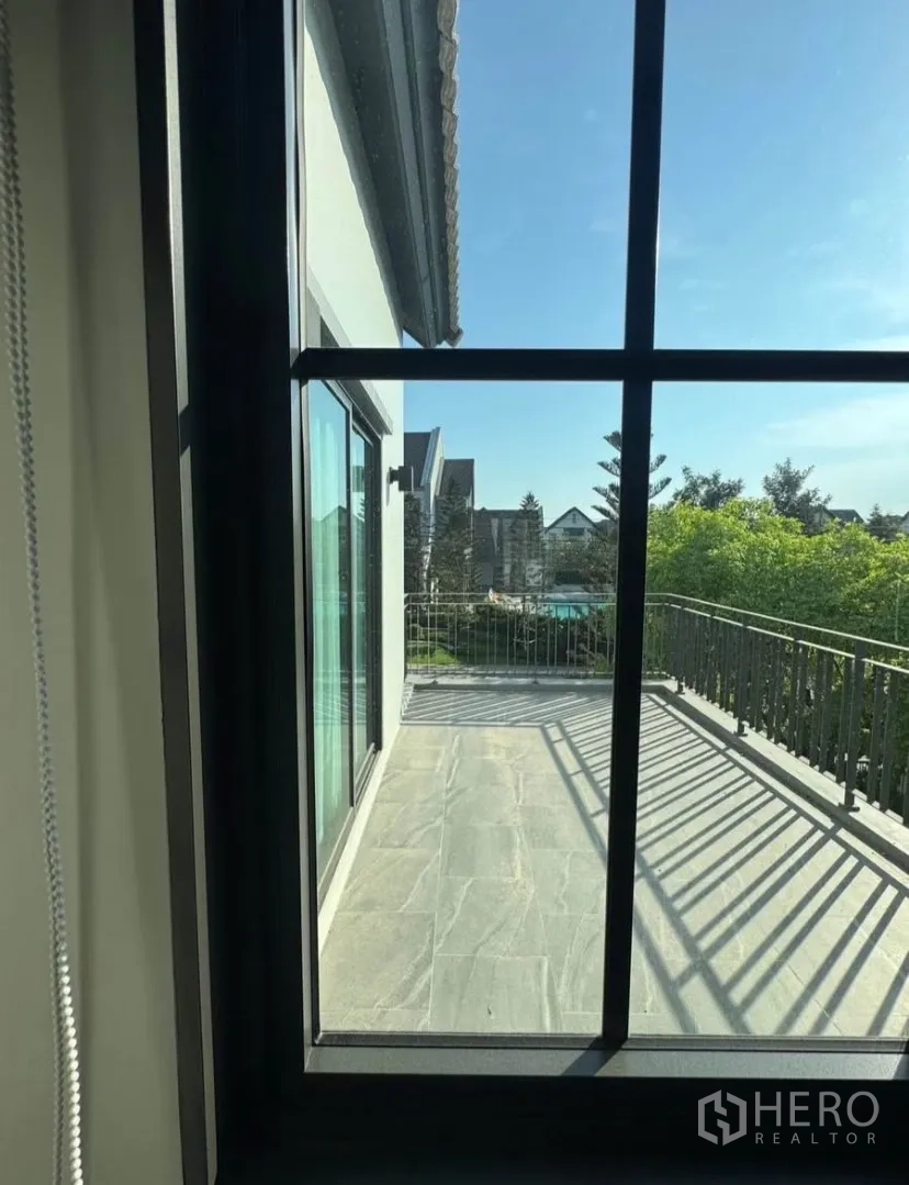 บ้านเดี่ยว for rent or sale in Lat Krabang, Bangkok - มุมมองผ่านหน้าต่างออกไปยังระเบียงชั้นบน ราวเหล็ก และต้นไม้ของหมู่บ้านใต้ท้องฟ้าสีฟ้า
