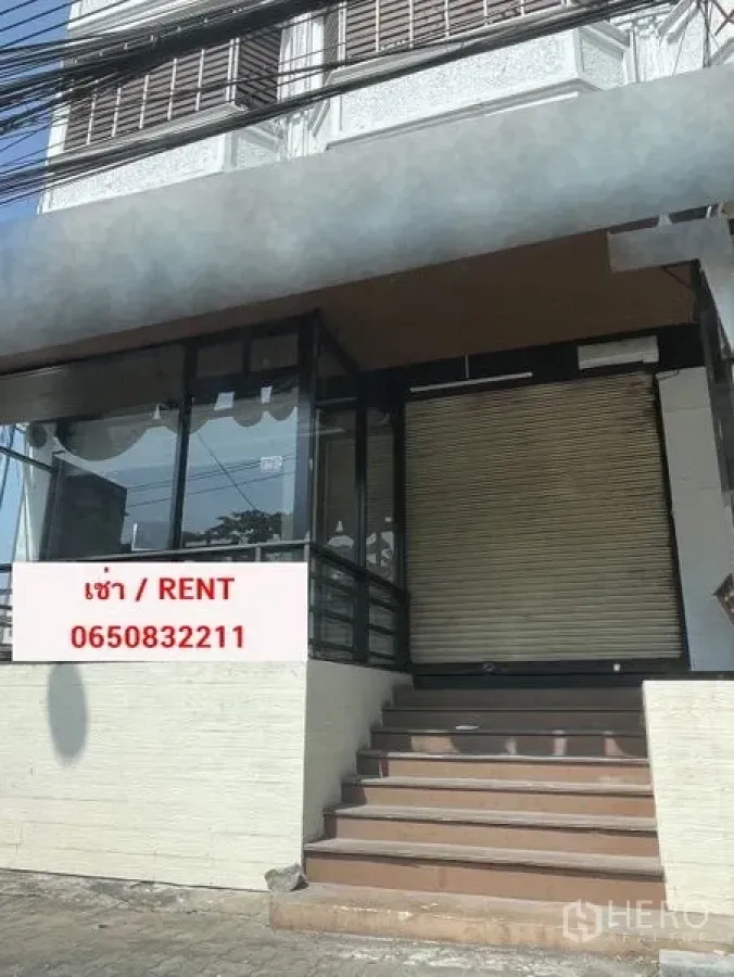 零售空间 出租在Bang Na, Bangkok - 邦那临街商铺外立面，整面玻璃、卷闸门、台阶入口与出租标识。