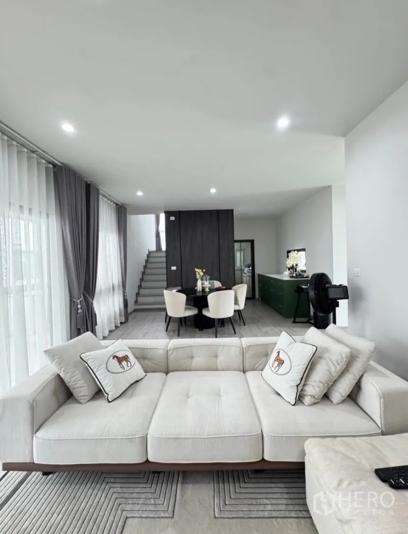 บ้านเดี่ยว for rent or sale in Lat Krabang, Bangkok - พื้นที่นั่งเล่นและรับประทานอาหารแบบโอเพ่นแพลน พร้อมหน้าต่างบานใหญ่ ผ้าม่านสีเทา และบันไดขึ้นชั้นบน