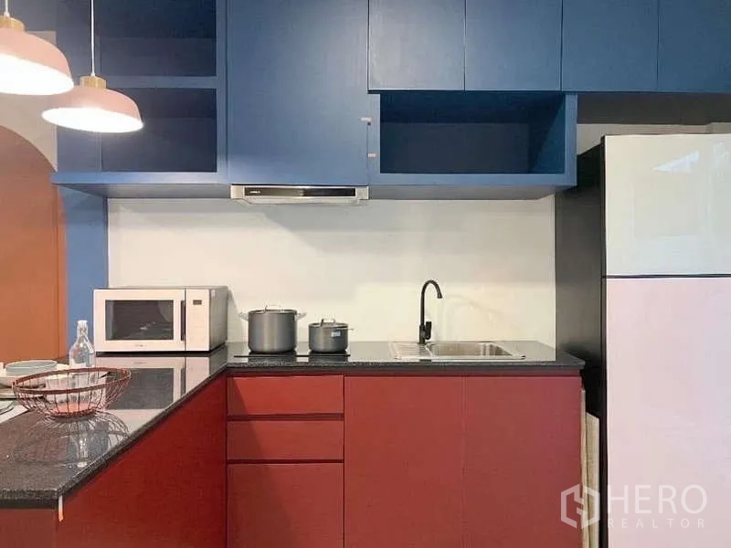 Townhouse for rent in Bang Bo, Samut Prakan - Modern L-shaped kitchen with blue upper cabinets, red base units, black granite countertop, sink and microwave.