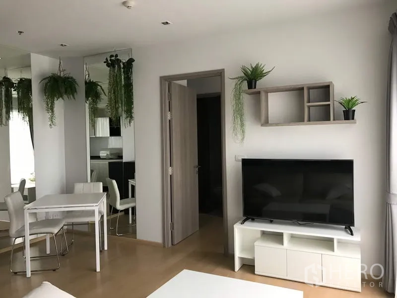 Condominium for rent in Watthana, Bangkok - Living area with flat-screen TV, wall shelves, and open doorway beside a compact dining set.
