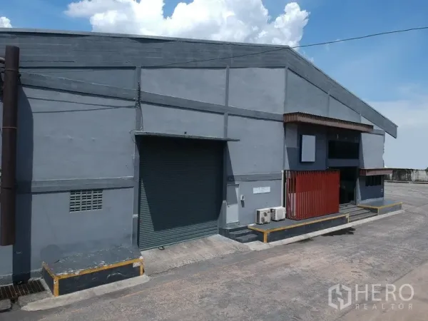 工厂 出租在Phra Pradaeng, Samut Prakan - 灰色厂房立面，带装卸平台、斜坡与卷闸门，天空晴朗。