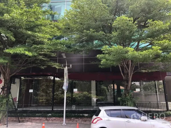 零售空间 出租在Watthana, Bangkok - 临街玻璃外立面配红色遮篷，门前有大树与停车位。