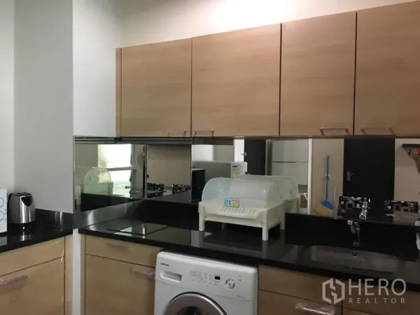 คอนโด ให้เช่าในPathum Wan, Bangkok - ครัวขนาดกะทัดรัด ตู้ไม้โทนอ่อน เคาน์เตอร์หินสีดำ ผนังกระจก และเครื่องซักผ้าฝาหน้า