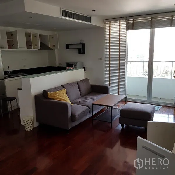 公寓 出租在Pathum Wan, Bangkok - 明亮客厅，灰色沙发与茶几，木地板，落地推拉门连通阳台，旁为开放式厨房。