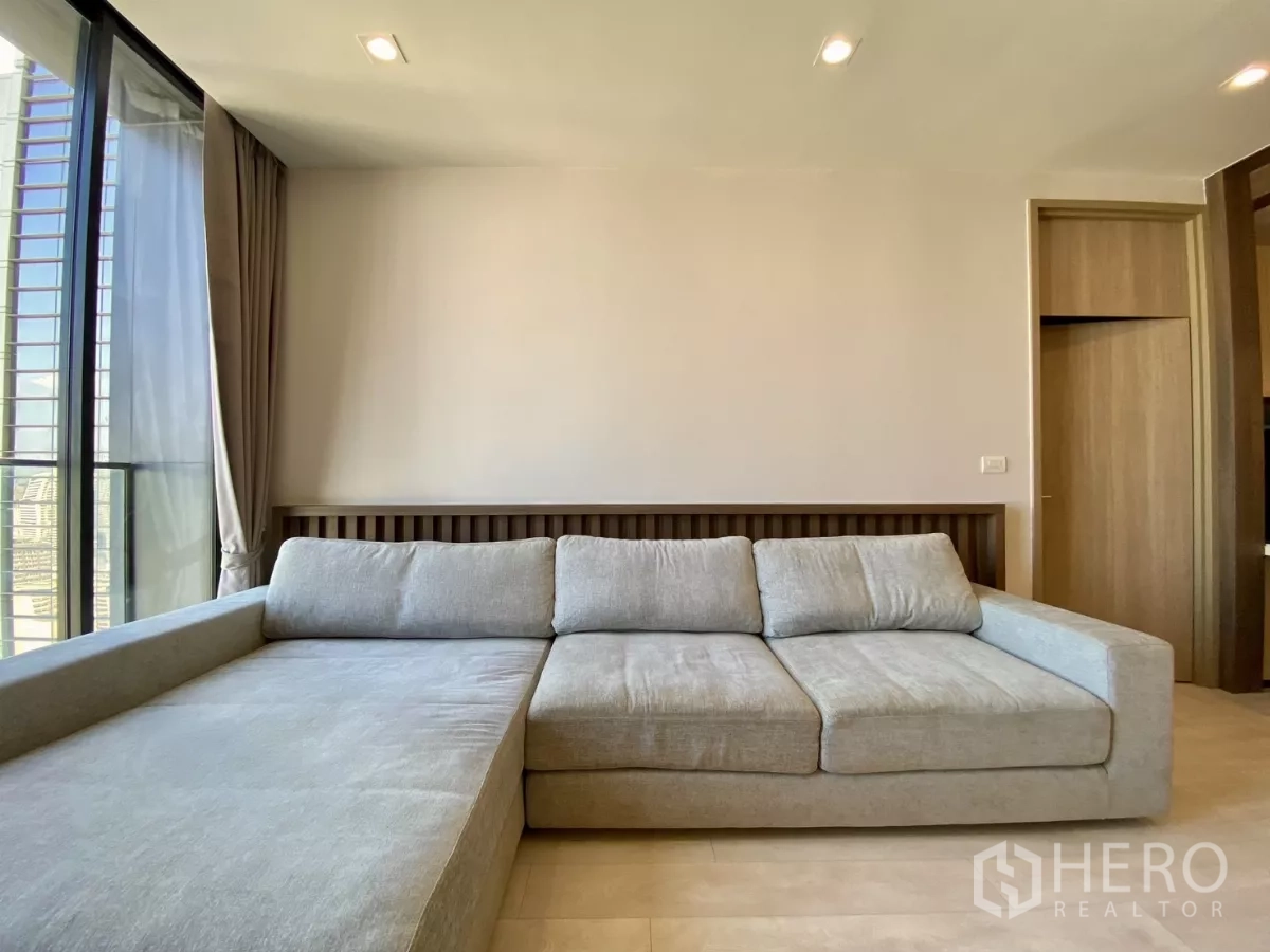 คอนโด ขายในPathum Wan, Bangkok - ห้องนั่งเล่นกว้าง โซฟา L ขนาดใหญ่ ผนังกระจกสูงพื้นจรดเพดาน โทนอบอุ่น