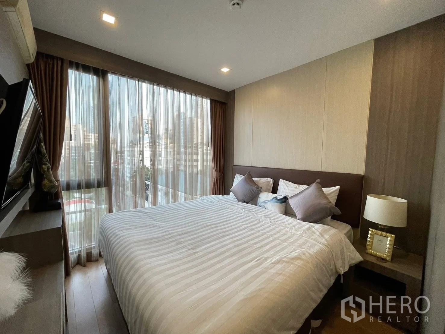 Condominium for rent in Watthana, Bangkok - Bedroom with a large bed, bedside table and lamp, built-in TV shelf, and city view through sheer curtains.