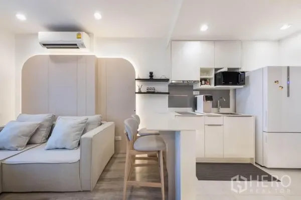 Condominium for rent in Bang Na, Bangkok - Compact kitchen with induction hob, sink, microwave, refrigerator, and a two-seat breakfast counter beside the sofa.