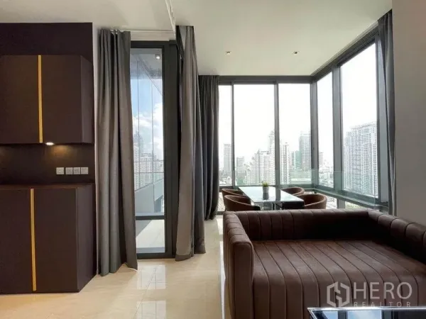Condominium for rent in Bang Rak, Bangkok - Bright living room with brown sofa, full-height windows and dining corner with city views.