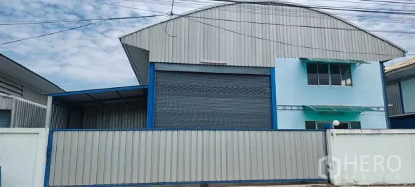 Warehouse for rent in Pathum Thani - Front view of gated warehouse with corrugated cladding, large shutter door, office windows and awnings beneath an arched roof in Pathum Thani.