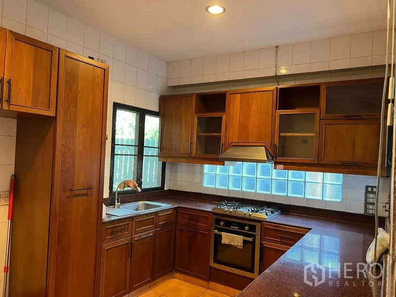Detached House for rent in Bang Phli, Samut Prakan - U-shaped kitchen with wooden cabinetry, gas hob, oven, stainless-steel sink and glass-block window.