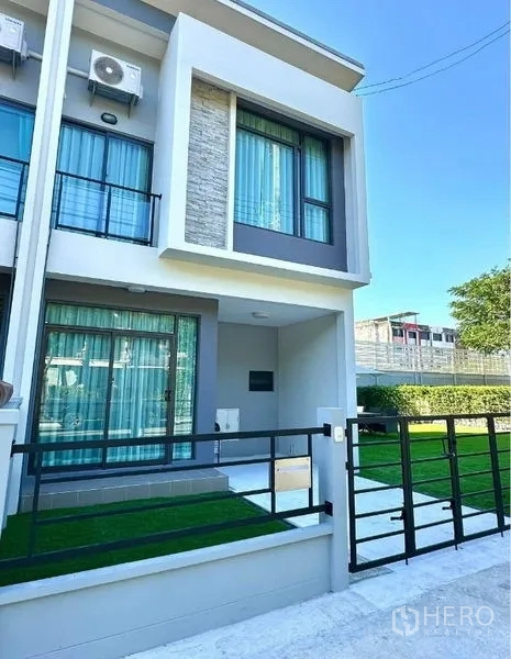 Townhouse for sale in Sai Mai, Bangkok - Modern two-storey townhouse facade with balcony, sliding glass doors, black gate, and small lawn in Bangkok.