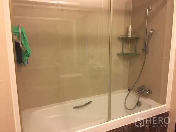 Condominium for rent in Watthana, Bangkok - Bathroom featuring a glass-enclosed bathtub with handheld shower and beige wall tiles.