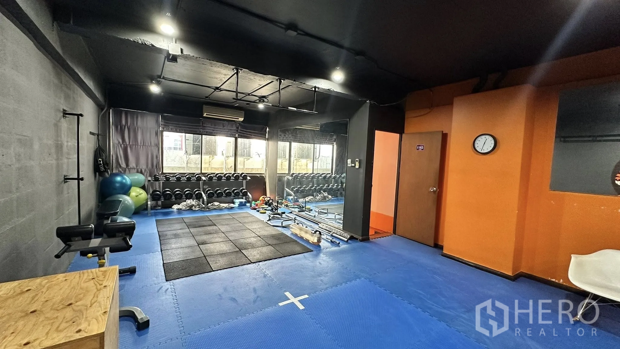 ออฟฟิศ for rent in Huai Khwang, Bangkok - มุมเวิร์กเอาต์มีชั้นวางดัมเบลริมหน้าต่าง พื้นยางสีน้ำเงิน และผนังสีส้ม