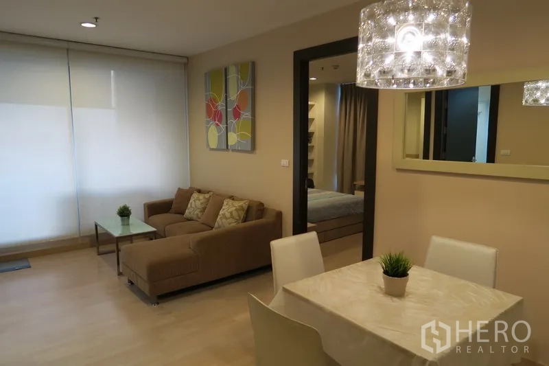 Condominium for rent in Huai Khwang, Bangkok - Bright living and dining area with L‑shaped sofa, glass coffee table, crystal light and large window, opening to a bedroom.