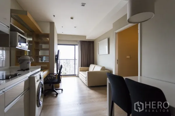 公寓 出租在Khlong Toei, Bangkok - 明亮客厅，配沙发、书桌、落地阳台门与开放式小厨房和洗衣机。