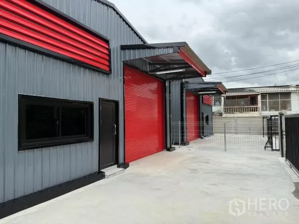 货栈 出租在Lat Phrao, Bangkok - 钢结构仓库侧视图，带行人门、窗户及多扇红色卷帘门。