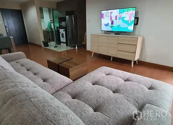 Condominium for rent in Huai Khwang, Bangkok - Sofa facing a TV on a light wood console with an open kitchen, fridge and washer in the background.
