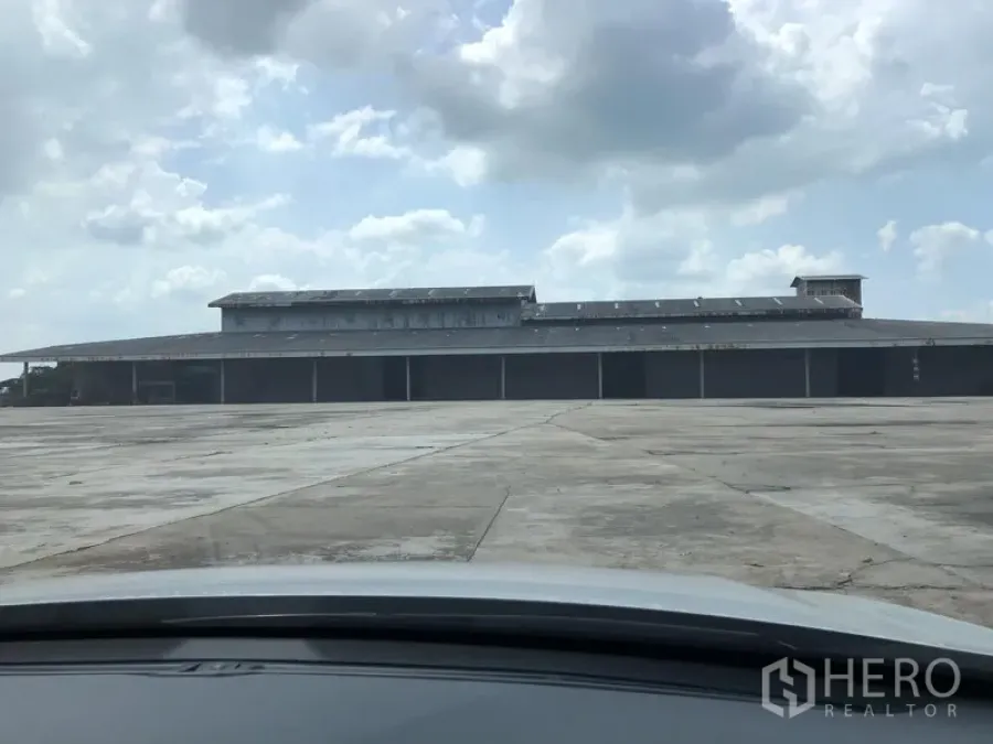 Warehouse for rent in Chaloem Phra Kiat, Saraburi - Large metal warehouse in Saraburi with multiple roller shutter bays and a vast concrete truck yard under a cloudy sky.