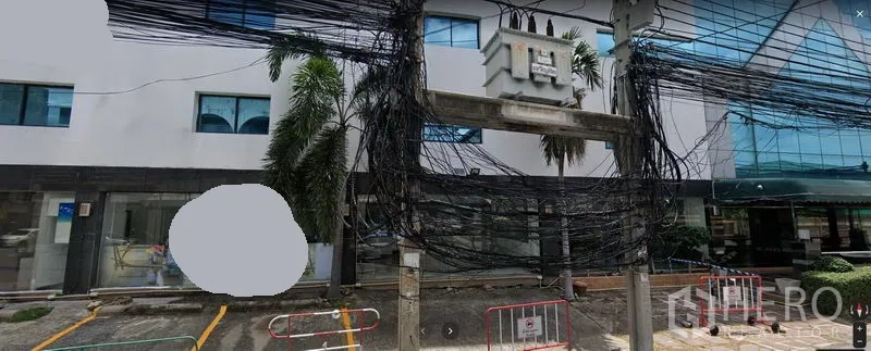 办公空间 出租在Watthana, Bangkok - 曼谷瓦他那区低层办公楼外观，玻璃立面、棕榈树、上方电缆与前方划线停车位。