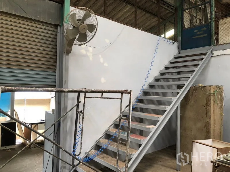 货栈 出租在Khlong Sam Wa, Bangkok - 仓库内的金属楼梯，旁有卷帘门与壁挂风扇。