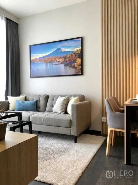 Condominium for rent in Watthana, Bangkok - Bright living room with grey sofa, soft rug and artwork, framed by a tall window with sheer curtains.