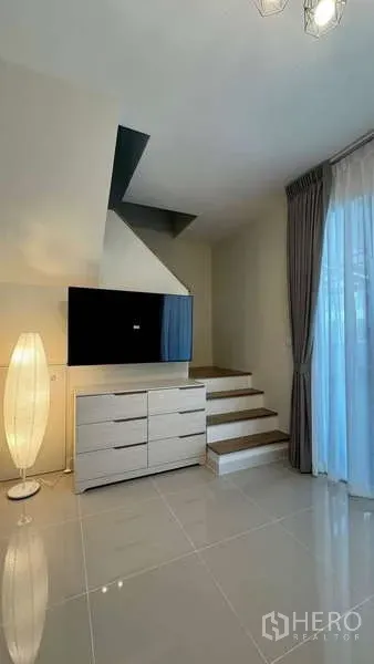 Townhouse for rent in Bang Phli, Samut Prakan - TV console set beneath staircase beside tall floor lamp and draped glass doors.