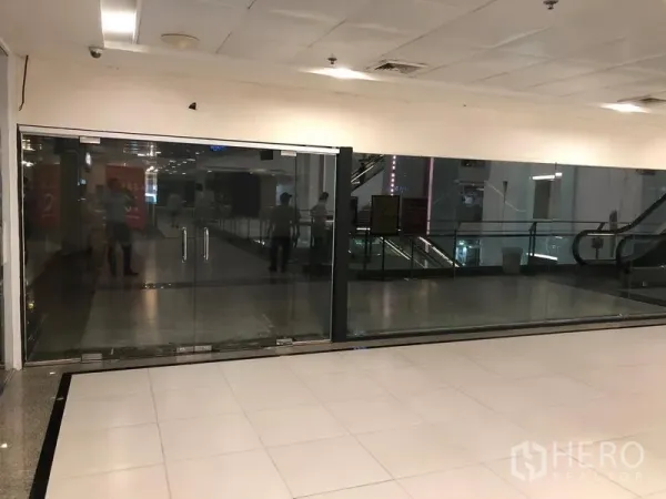 零售空间 出租在Watthana, Bangkok - 购物中心走道旁的空置玻璃门商铺，地砖地面，旁有自动扶梯