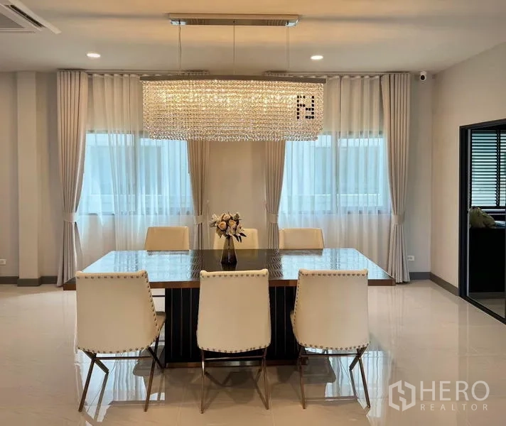 Detached House for rent or sale in Bang Phli, Samut Prakan - Dining room with six chairs, a large crystal chandelier, and wide windows with curtains.