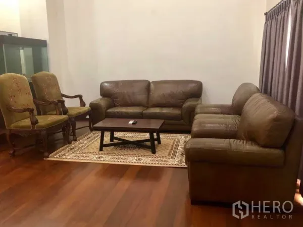 Townhouse for rent in Khlong Toei, Bangkok - Spacious living room featuring brown leather sofas, classic armchairs, a coffee table, and a patterned rug.