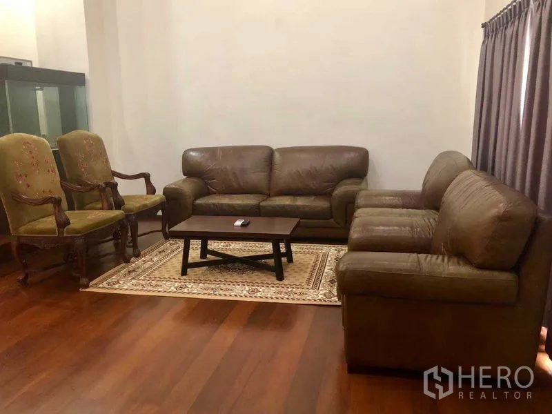 Townhouse for rent in Khlong Toei, Bangkok - Spacious living room featuring brown leather sofas, classic armchairs, a coffee table, and a patterned rug.