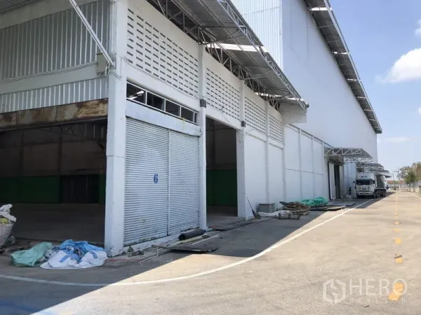 货栈 出租在Pathum Thani - 仓库外立面设多扇卷帘门、带雨棚装卸区及宽阔卡车车道（巴吞他尼）。