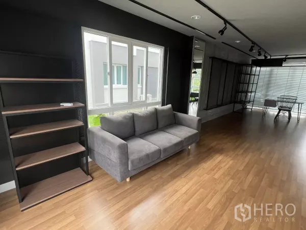 Detached House for rent or sale in Don Mueang, Bangkok - Seating corner with grey sofa by bright windows and shelving unit.