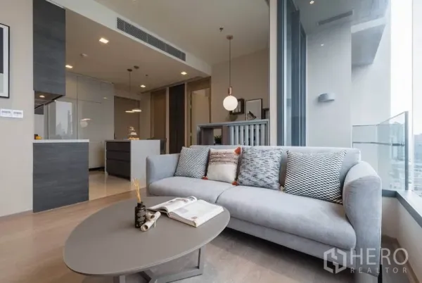 Condominium for rent in Watthana, Bangkok - Bright living room with gray sofa, coffee table, and large windows facing city views next to the kitchen.