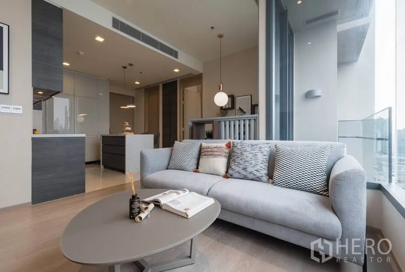Condominium for rent in Watthana, Bangkok - Bright living room with gray sofa, coffee table, and large windows facing city views next to the kitchen.