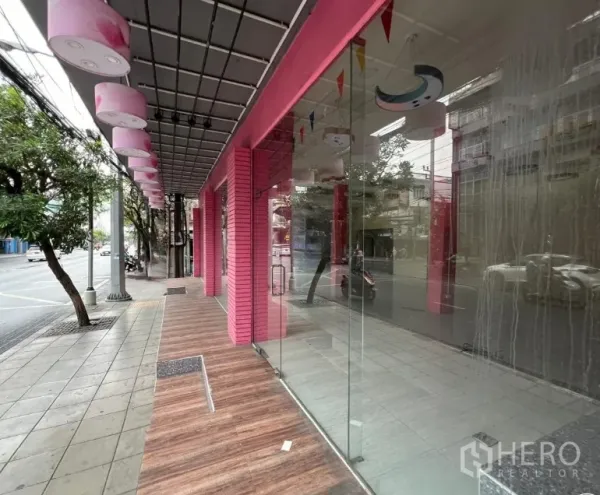 店屋 出租在Watthana, Bangkok - 靠树荫街道的一侧视角，带顶走道与大面积玻璃门面。