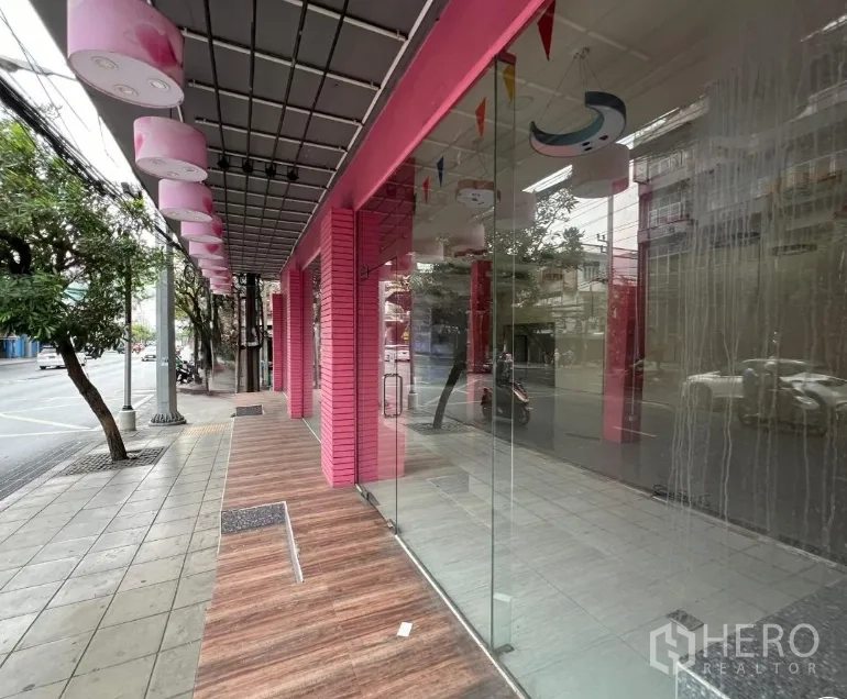 店屋 出租在Watthana, Bangkok - 靠树荫街道的一侧视角，带顶走道与大面积玻璃门面。