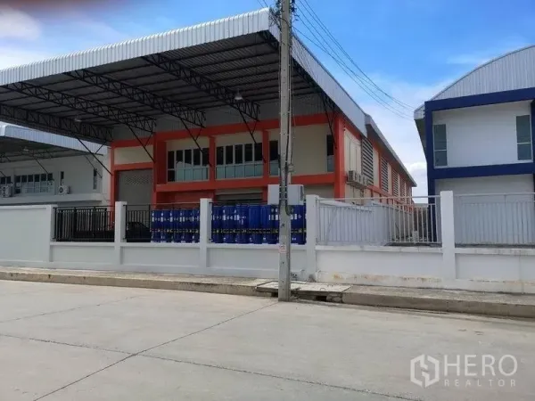 Factory for rent in Samut Prakan - Modern factory exterior with covered loading bay, roller shutter and gated yard on a wide concrete road.