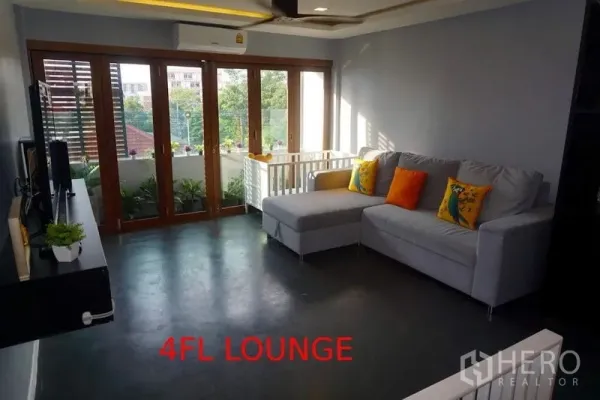 店屋 出售在Phra Khanong, Bangkok - 采光充足的客厅，L形沙发、电视墙，落地玻璃门通向阳台。