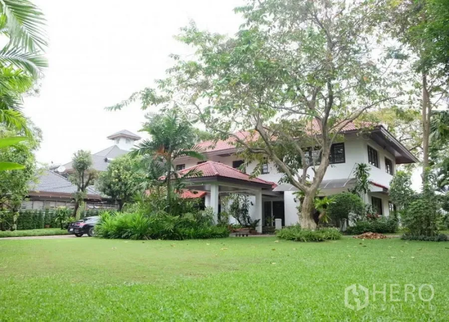 独立式住宅 出租在Bang Phli, Samut Prakan - 社区内大草坪与成树环绕的两层独栋别墅，红色坡屋顶。