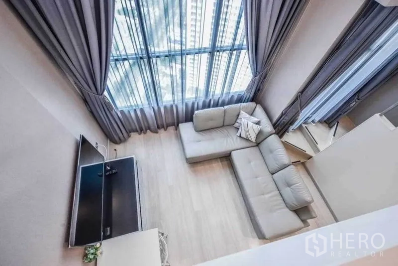 Condominium for rent in Sathon, Bangkok - Aerial view of living area showing sectional sofa, TV stand and tall windows with drapes.