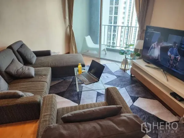 Condominium for rent in Huai Khwang, Bangkok - Living area with sectional sofa facing TV and sliding glass doors to a balcony with city view.